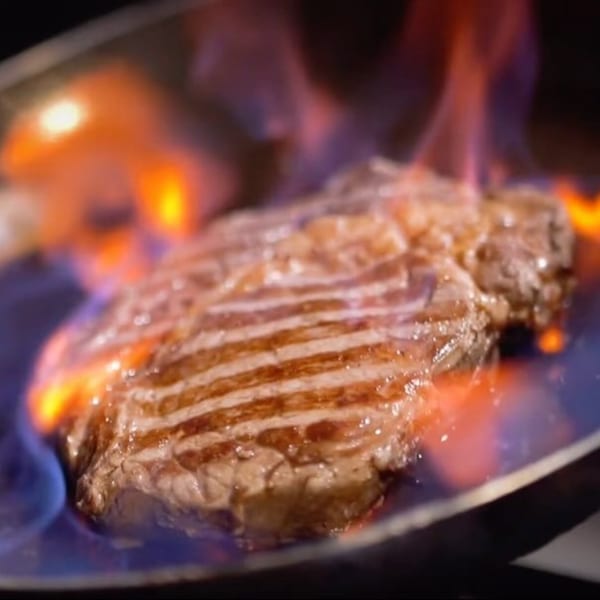 A thick steak with grill marks is flambéed in a pan, surrounded by bright orange and blue flames.