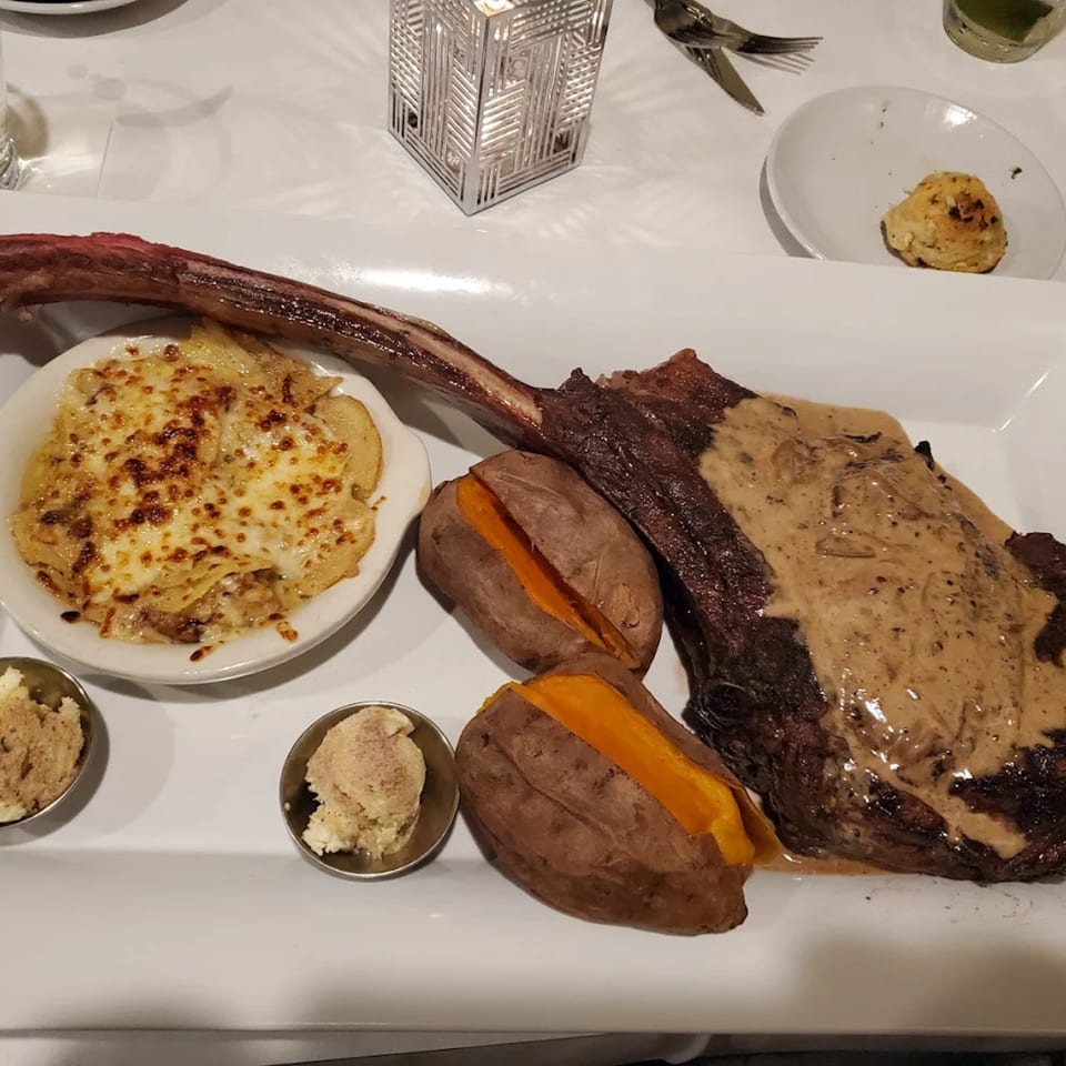 A large tomahawk steak with sauce, two baked sweet potatoes, and cheesy scalloped potatoes.